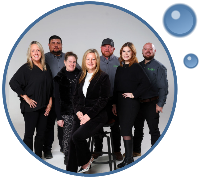 Seven adults pose together for a group photo in a studio, with three seated and four standing, all dressed in business or casual attire. A blue circular border and bubbles frame the image.